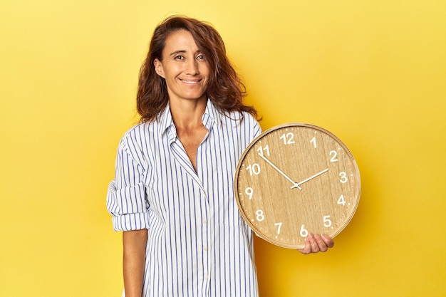 Frau mittleren Alters hält eine Wanduhr vor gelbem Hintergrund, glücklich lächelnd und fröhlich