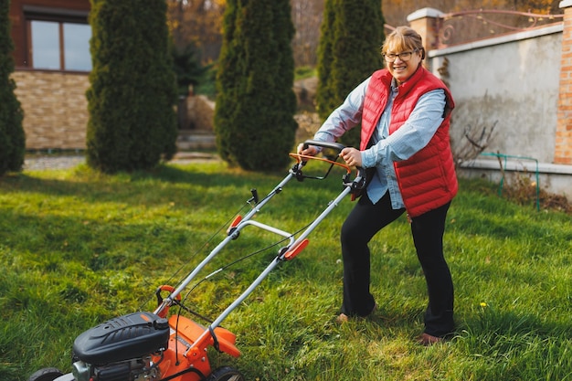 Frau mittleren Alters in roter Weste mit Rasenmäher auf Hinterhof, Blick in die Kamera Gärtnerin, die im Sommer oder Herbst arbeitet und Gras im Hinterhof schneidet Konzept der Gartenarbeit Natur