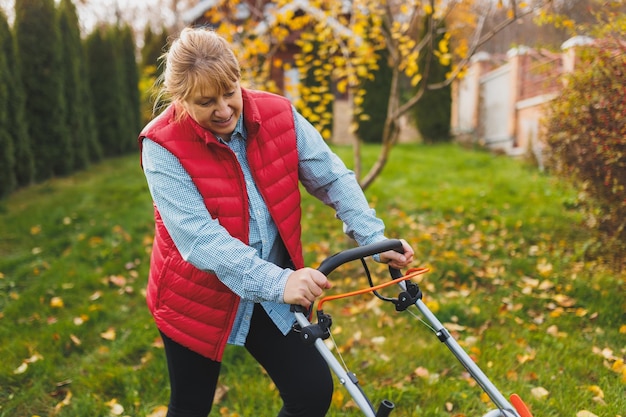 Frau mittleren Alters in roter Weste mit Rasenmäher auf Hinterhof, Blick in die Kamera Gärtnerin, die im Sommer oder Herbst arbeitet und Gras im Hinterhof schneidet Konzept der Gartenarbeit Natur