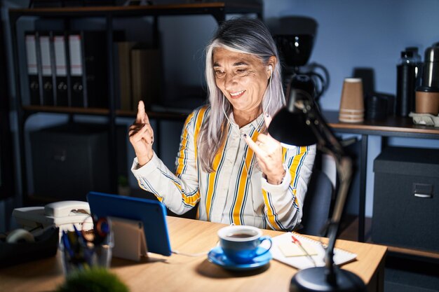 Foto frau mittleren alters mit grauen haaren, die nachts im büro arbeitet und mit verrücktem gesichtsausdruck schreit und mit erhobenen händen rocksymbole macht. musikstar. schweres konzept.