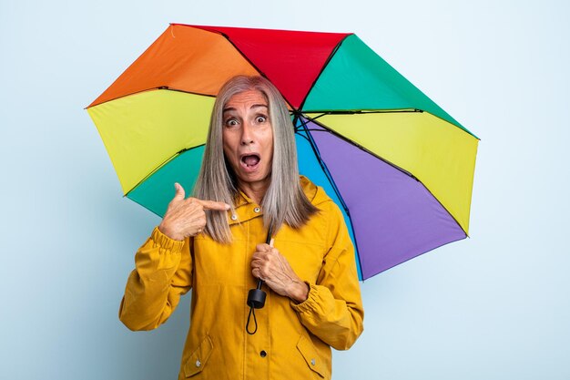 Frau mittleren Alters mit grauen Haaren, die schockiert und überrascht aussieht, mit weit offenem Mund, der auf das Konzept des Regenschirms und des Regens zeigt