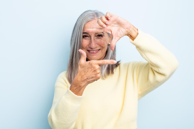 Frau mittleren Alters mit grauen Haaren, die sich glücklich, freundlich und positiv fühlt, lächelt und ein Porträt oder einen Fotorahmen mit den Händen macht