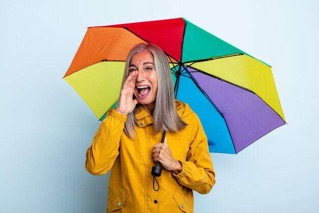 Frau mittleren Alters mit grauen Haaren, die sich glücklich fühlt und mit den Händen neben dem Mund einen großen Schrei ausspricht. Regenschirm und Regenkonzept