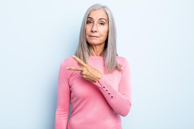 Frau mittleren Alters mit grauen Haaren, die sich glücklich, positiv und erfolgreich fühlt, mit Hand, die V-Form über der Brust macht und Sieg oder Frieden zeigt