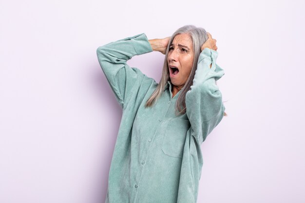 Frau mittleren Alters mit grauen Haaren mit offenem Mund, die wegen eines schrecklichen Fehlers entsetzt und schockiert aussieht und die Hände zum Kopf hebt