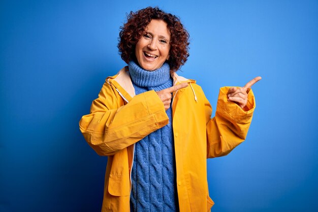 Frau mittleren Alters mit lockigem Haar und Regenmantel, die über isoliertem blauem Hintergrund steht, lächelt und in die Kamera schaut, die mit zwei Händen und Fingern zur Seite zeigt