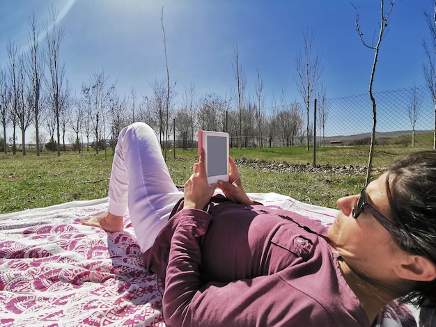 Frau mittleren Alters mit Sonnenbrille, die an einem schönen Tag ein E-Book liest, das auf dem Gras liegt