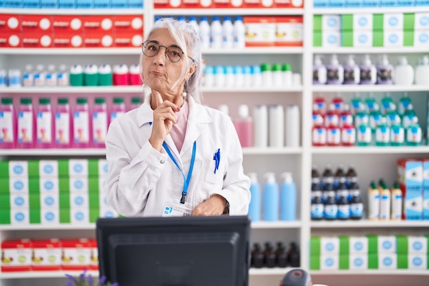 Frau mittleren Alters mit Tätowierungen, die in einer Apotheke arbeitet, denkt konzentriert über Zweifel nach, den Finger am Kinn und schaut verwundert nach oben