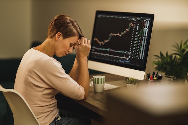 Frau mittleren Alters müde, erschöpft von der Arbeit am Tisch und dem Handel mit Kryptowährung spät in der Nacht im Heimbüro.