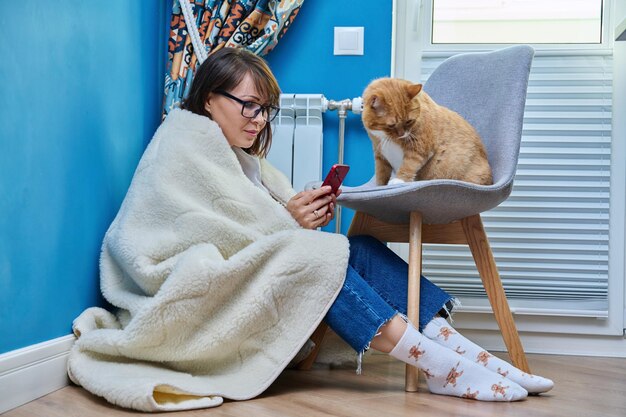 Frau mittleren Alters ruht sich zu Hause unter einer warmen Wolldecke aus und nutzt ihr Smartphone mit einer Katze in der Nähe eines Heizkörpers. Winterherbst Heizsaison Lifestyle Freizeit sparen Energiekrisenkonzept