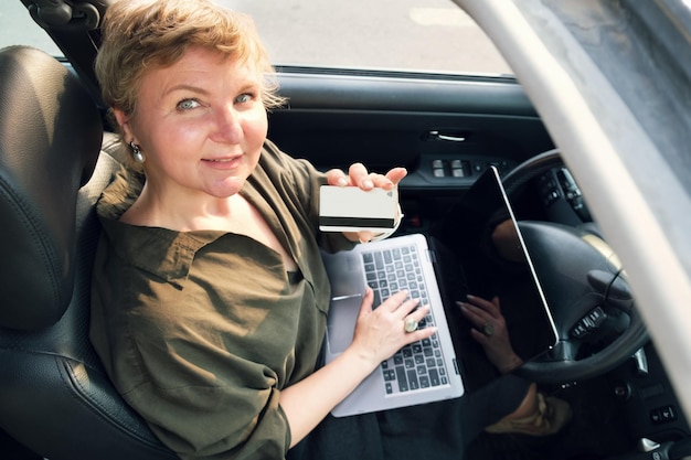 Frau mittleren Alters sitzt hinter dem Steuer eines Autos mit einem Laptop auf ihrem Schoß