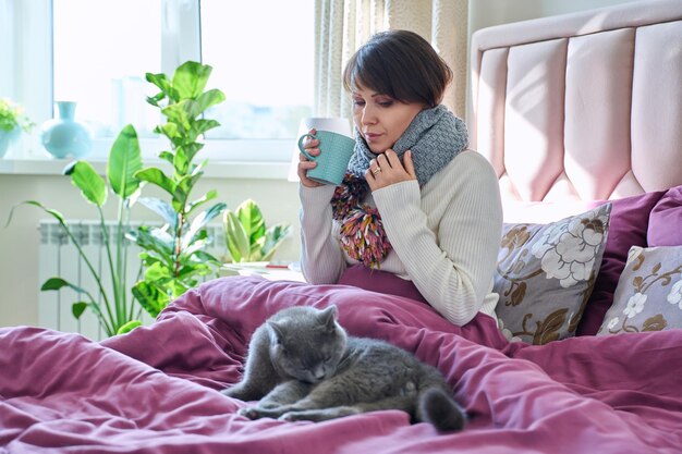 Frau mittleren Alters sitzt zu Hause im Bett und wärmt sich mit einem Schal und einem Becher Heißgetränk zusammen mit einer grauen Katze. Lifestyle-Konzept für gemütliche kalte Jahreszeit, Herbst, Winter, Komfort, Tiere, Menschen