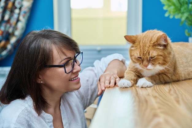 Frau mittleren Alters spricht mit rothaariger Hauskatze im Hintergrund