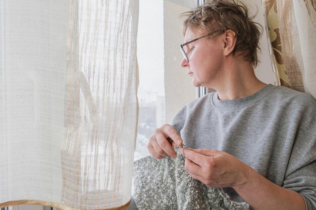 Frau mittleren Alters strickt einen grauen Wollschal, der am Fenster sitzt Die Frau macht Handarbeiten