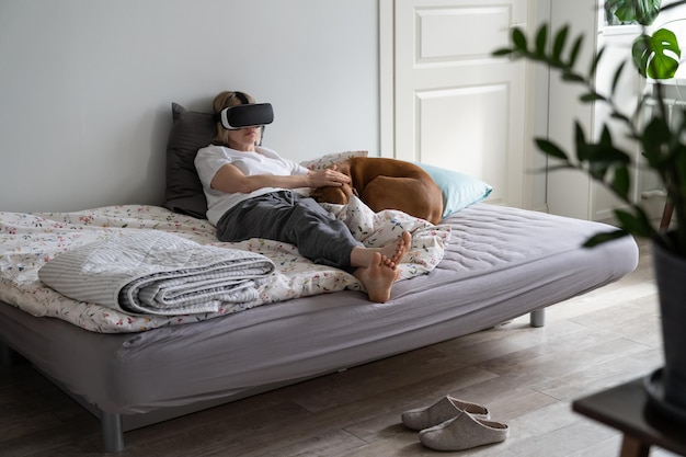 Frau mittleren Alters verbringt ihre Freizeit damit, VR-Spiele zu spielen, die auf dem Bett neben schlafendem Hund liegen