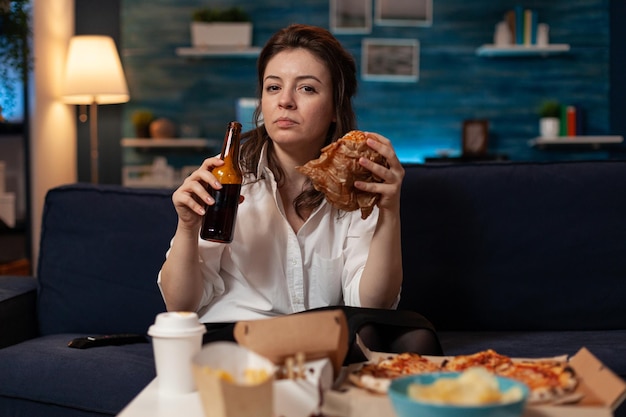 Frau nach der Büroarbeit, die leckeren Hamburger zum Mitnehmen isst und Bierflasche auf der Couch hält und Nachrichten im Fernsehen sieht. Ernsthafte Person mit Burger zum Mitnehmen und Fast-Food-Liefermenü.