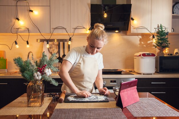 Frau nach Rezept auf digitaler Tablette und knetet den Teig zum Kochen von Weihnachtslebkuchen in ihrer Küche.