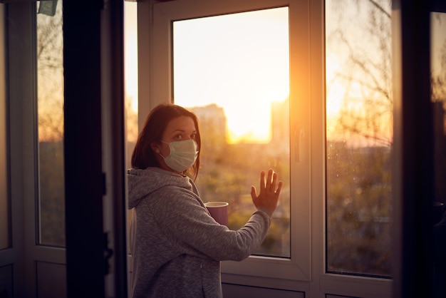 Frau nahe Fenster bei Sonnenuntergang isoliert zu Hause für Virusausbruch. Bleib zu Hause Konzept