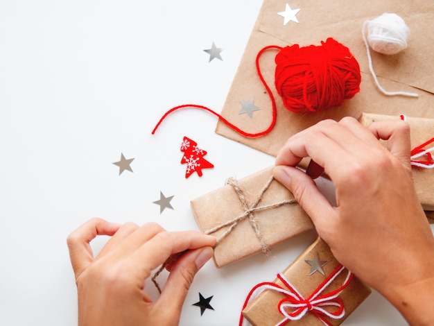 Frau packt Weihnachts- und Neujahrs-DIY-Geschenke in Kraftpapier. Geschenke mit weißen und roten Fäden gebunden.