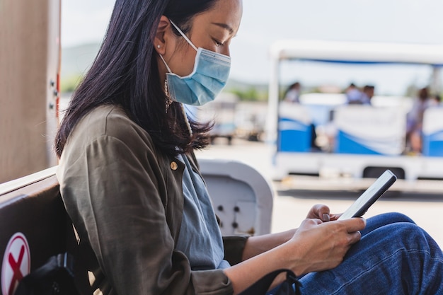 Frau Passagier in der medizinischen Maske unter Verwendung des Smartphones beim Sitzen in Flughafentransporten.