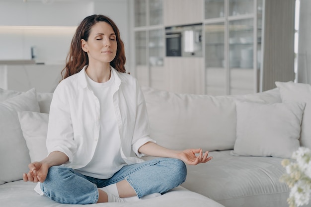 Frau praktiziert Yoga sitzt auf dem Sofa in Lotus-Pose Gesunde Gewohnheiten achtsamer Lebensstil Angstlinderung