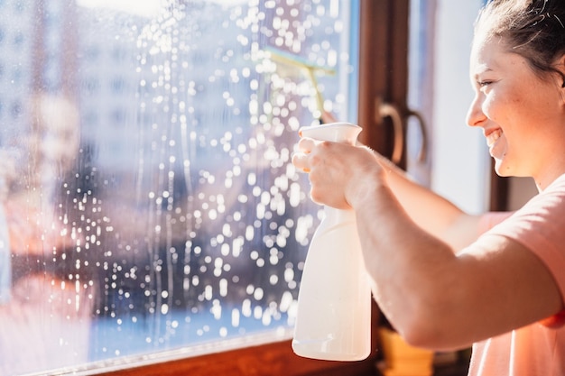 Frau putzt ein Fenster mit Reinigungssprüher Housekeeping-Konzept Mädchen halten Reinigungsrakel in der Hand