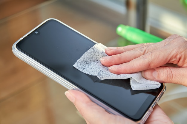 Frau putzt ein Smartphone mit einem Strahl Alkohol Aerosol Händedesinfektionsmittel und Einweg-Tuch zu Hause. Vorsichtsmaßnahmen bezüglich Viren