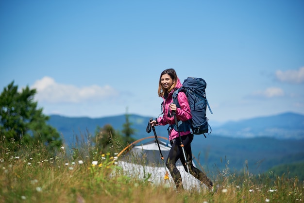 Frau Rucksacktourist mit Rucksack und Trekkingstöcken in der Nähe von Zelt, auf der Spitze eines Hügels gegen blauen Himmel gehen, sonnigen Tag in den Bergen genießen.