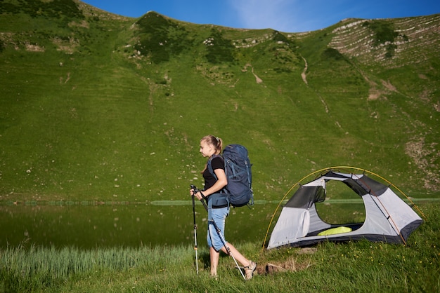 Frau Rucksacktourist mit Rucksack und Trekkingstöcken nahe Zelt, Sommertag nahe dem See in den Bergen genießend