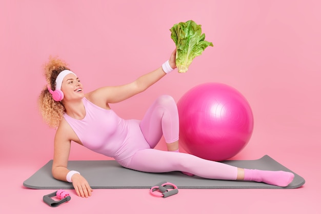 Frau schaut auf grünes Gemüse motiviert Sie zu einem gesunden Lebensstil in Activewear gekleidet liegt auf Fitnessmatte hört Musik über Kopfhörer macht Pause nach langem Training