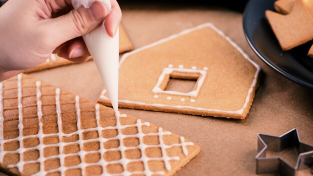 Frau schmückt Lebkuchenhaus mit weißem Zuckerguss auf Holztischhintergrund, Backpapier in der Küche, Nahaufnahme, Makro.