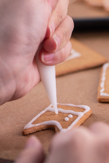 Frau schmückt Lebkuchenhaus mit weißem Zuckerguss auf Holztischhintergrund, Backpapier in der Küche, Nahaufnahme, Makro.