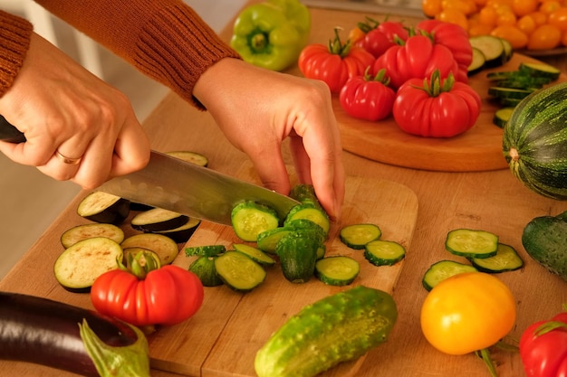 Frau schneidet mit einem Messer frisches rohes Gemüse, Tomaten und Gurken werden auf einem Holzbrett in Scheiben geschnitten. Herd zu Hause, Zubereitung einer gesunden vegetarischen Mahlzeit. Hacken von Zutaten für die Zubereitung eines vegetarischen Gerichts. 4K