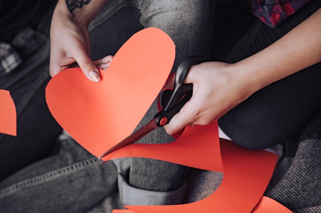 Frau schneidet rotes Papierherz mit einer Schere am Valentinstag auf dem Bett. Nahansicht