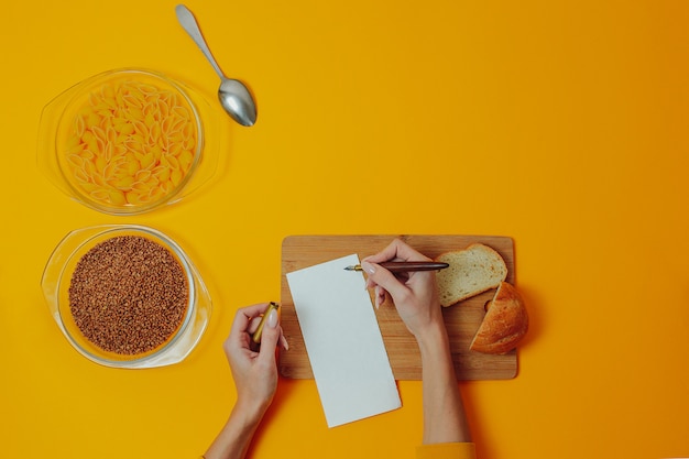 Frau schreibt Diätschritte oder -rezept auf das weiße Blatt. Buchweizen- und Makkaroniprodukte in den Glasplatten. Langsame Kohlenhydrate mit Kopienraum.