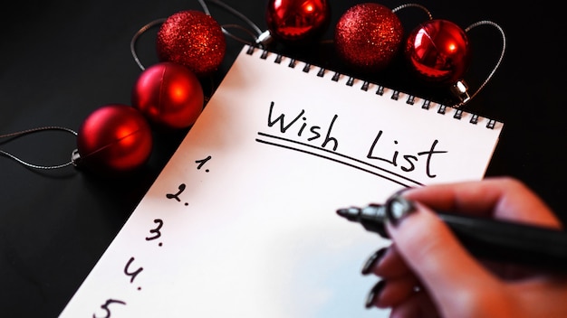 Frau schreibt ihre Wunschliste. Notizbuch auf schwarzem Hintergrund mit Weihnachtskugeln - Wunschliste des neuen Jahres
