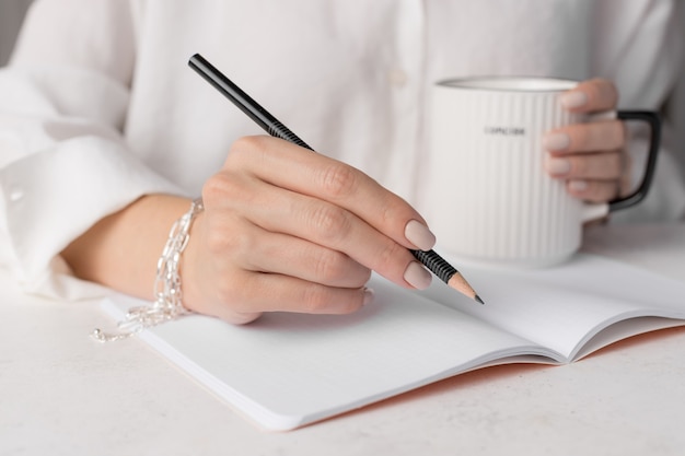 Frau schreibt in Notizblock auf weißem Schreibtisch mit Kaffeetasse Home-Office-Konzept Nahaufnahme