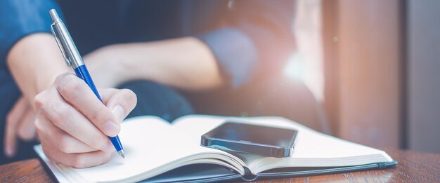 Foto frau schreibt mit einem blauen stift im büro handschriftlich auf einen notizblock. auf dem tisch liegt ein mobiltelefon