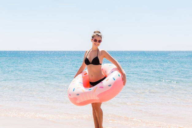 Frau schwimmt mit aufblasbarem Donut am Strand im sonnigen Sommertag. Sommerferien und Urlaubskonzept.