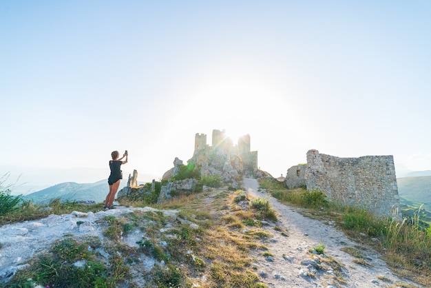 Frau selfie an Burgruine auf Berggipfel bei Rocca Calascio, italienisches Reiseziel, Wahrzeichen im Nationalpark Gran Sasso, Abruzzen, Italien. Die Sonne des klaren blauen Himmels platzte im Gegenlicht