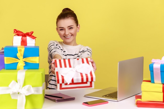 Frau sitzt am Arbeitsplatz mit Laptop und hält eine Geschenkbox in der Hand, die der Kamera gratuliert