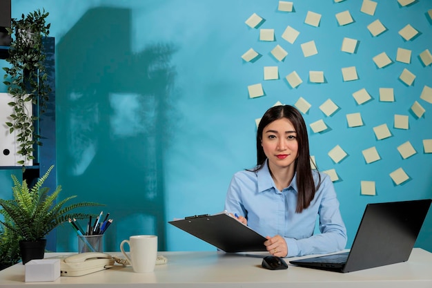 Frau sitzt am Schreibtisch und macht sich Notizen über den Nachschubbestand an Büromaterial. Mitarbeiterin des Unternehmens, die für die Einkaufsabteilung mit Laptop und Kaffeetasse auf dem Schreibtisch verantwortlich ist.