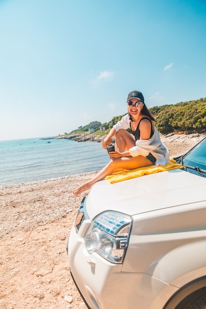 Frau sitzt an der Motorhaube und trinkt Kaffee mit Blick auf den Sommerurlaub am Meer