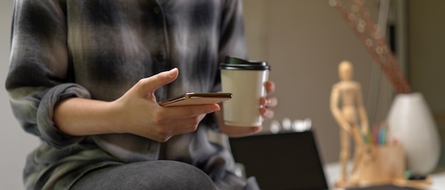 Frau sitzt auf Arbeitstisch und benutzt Smartphone, während Kaffee gehalten wird