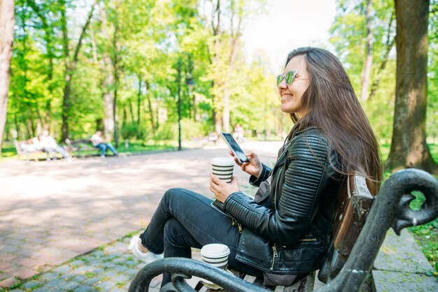 Frau sitzt auf Bank im Stadtpark, trinkt Kaffee und surft per Handy im Internet