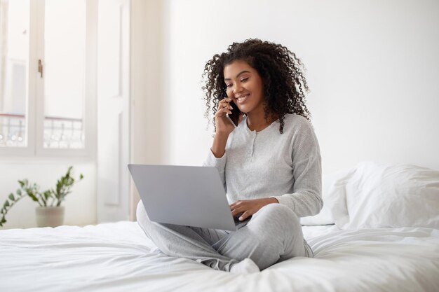 Frau sitzt auf dem Bett, spricht am Telefon und benutzt einen Laptop
