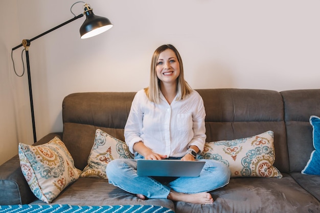 Frau sitzt auf der Couch im Wohnzimmer mit Laptop, surft im Internet und arbeitet fern von zu Hause aus