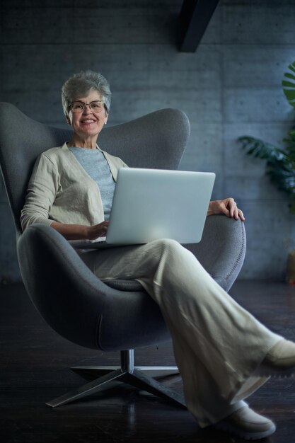 Frau sitzt auf einem modernen Stuhl am Fenster in einem hellen, gemütlichen Raum zu Hause Sie arbeitet in entspannter Atmosphäre am Laptop