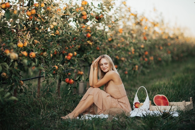 Frau sitzt auf einer weißen Decke draußen im Apfelgarten. Glückliche Frau, die Picknick im Herbstgarten mit Wassermelone, Äpfeln und Trauben hat.