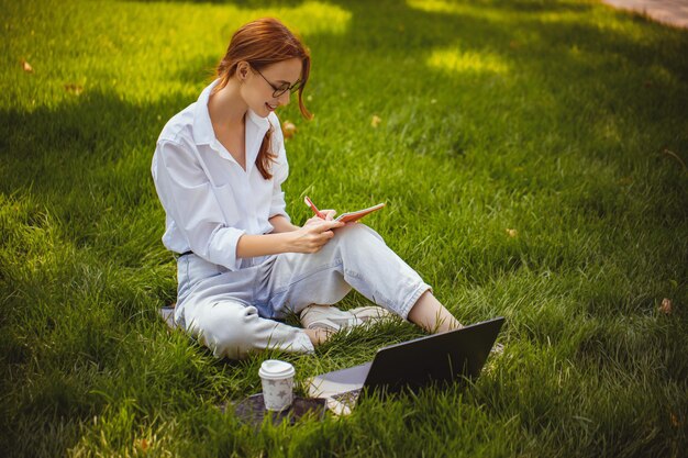 Frau sitzt auf Gras im Park und arbeitet am Laptop-PC und macht einige Notizen Freiberufler, der im Freien arbeitet